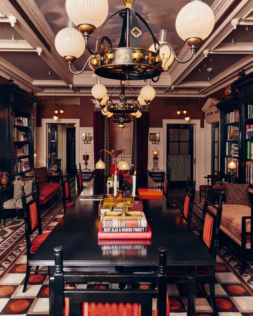 Lobby biblioteca de hotéis boutique em londres com mesa longa preta, estantes de livros e lustres dourados.