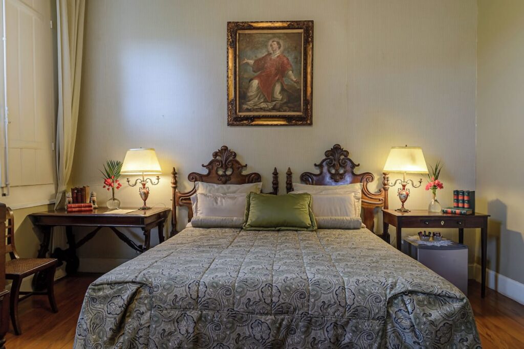 Quarto clássico com cabeceiras entalhadas e arte sacra, experiência autêntica em fazendas históricas para se hospedar.