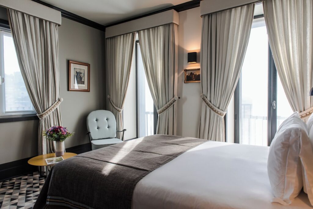 Quarto clássico do Valverde Hotel com cortinas beges, poltrona retrô e piso geométrico, destaque de design entre os hoteis boutique em lisboa.