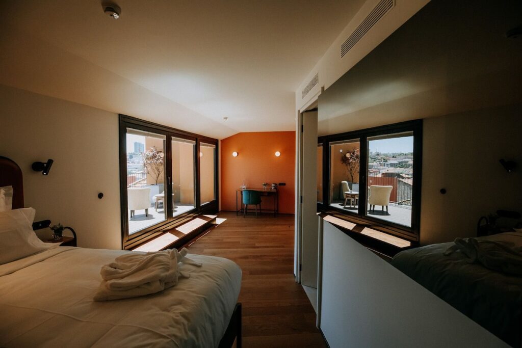 Quarto premium de hotel boutique no Porto com ampla varanda integrada por portas de vidro, piso de madeira acolhedor e parede de destaque terracota.