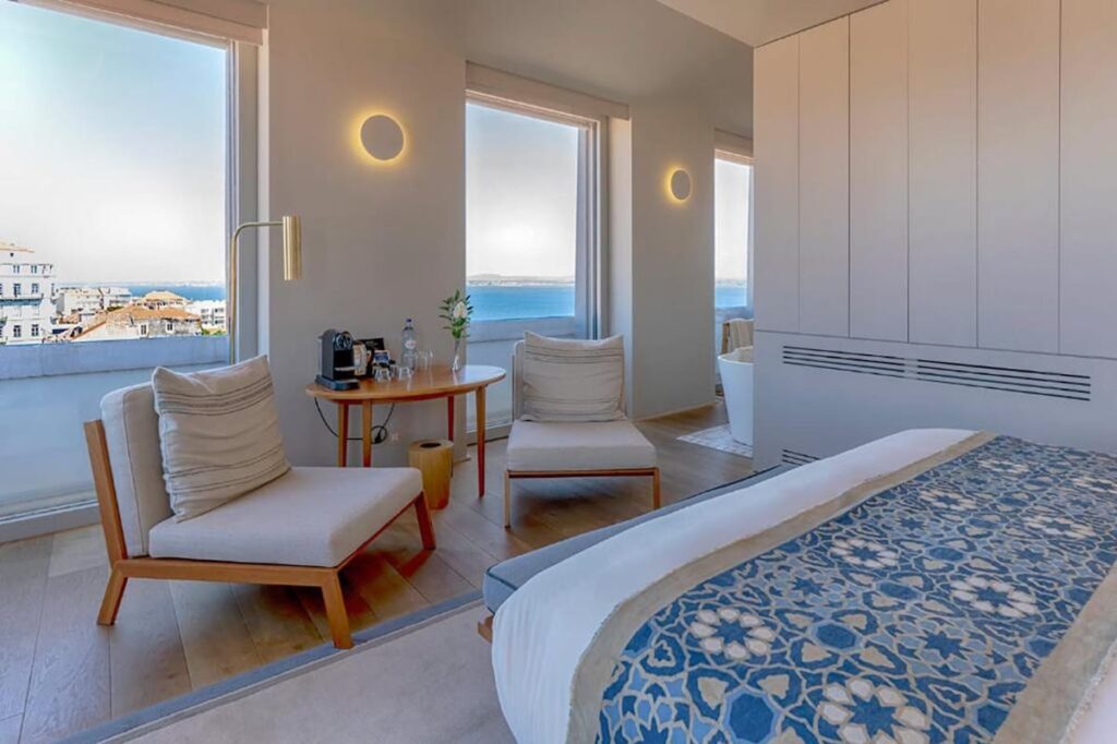 Suíte moderna no Verride Palácio Santa Catarina com vista do rio, poltronas de madeira e manta azul, requinte nos hoteis boutique em lisboa.