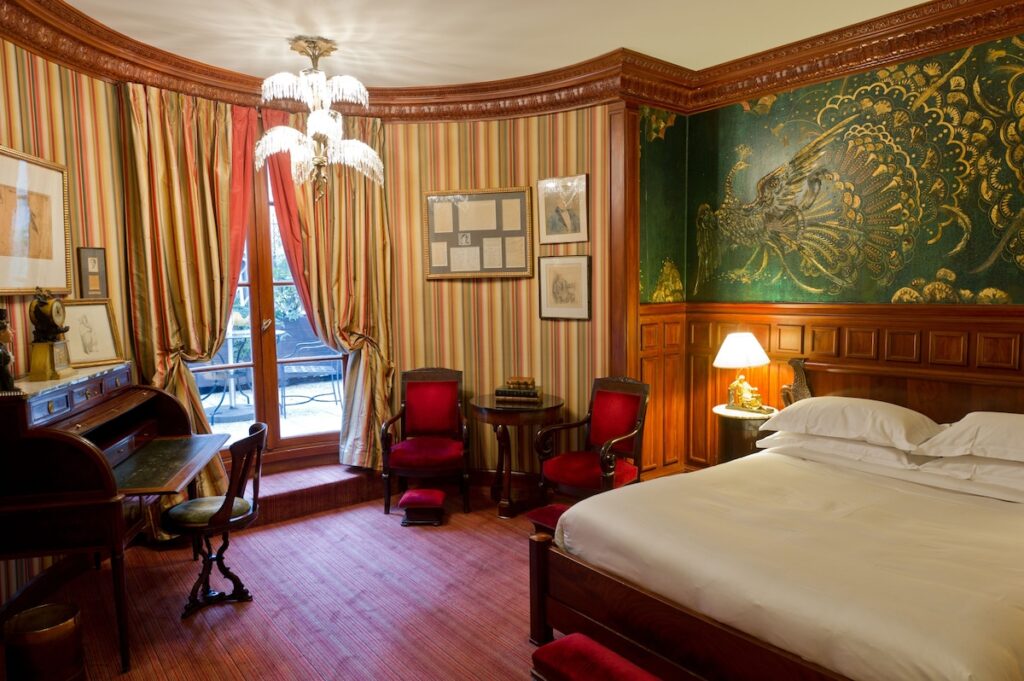 Quarto clássico do L'Hotel, um dos mais icônicos hotéis boutique em Paris, com cabeceira de pavão dourado, móveis de madeira nobre e decoração opulenta.