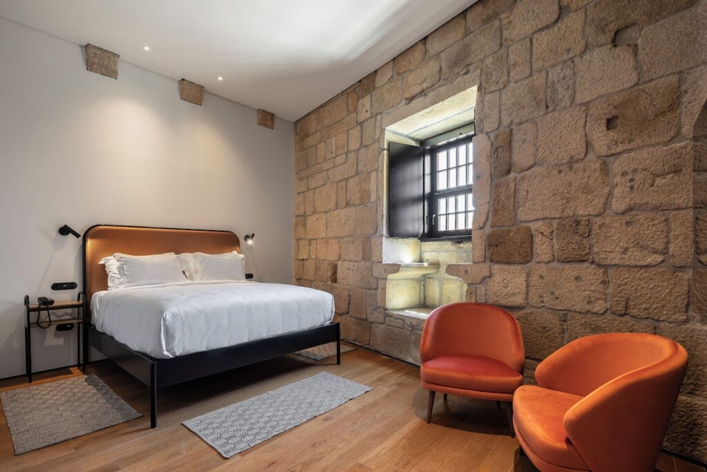 Acolhedor quarto superior de hotel boutique no Porto com parede original em pedra granítica exposta, duas poltronas em veludo laranja e piso de madeira.