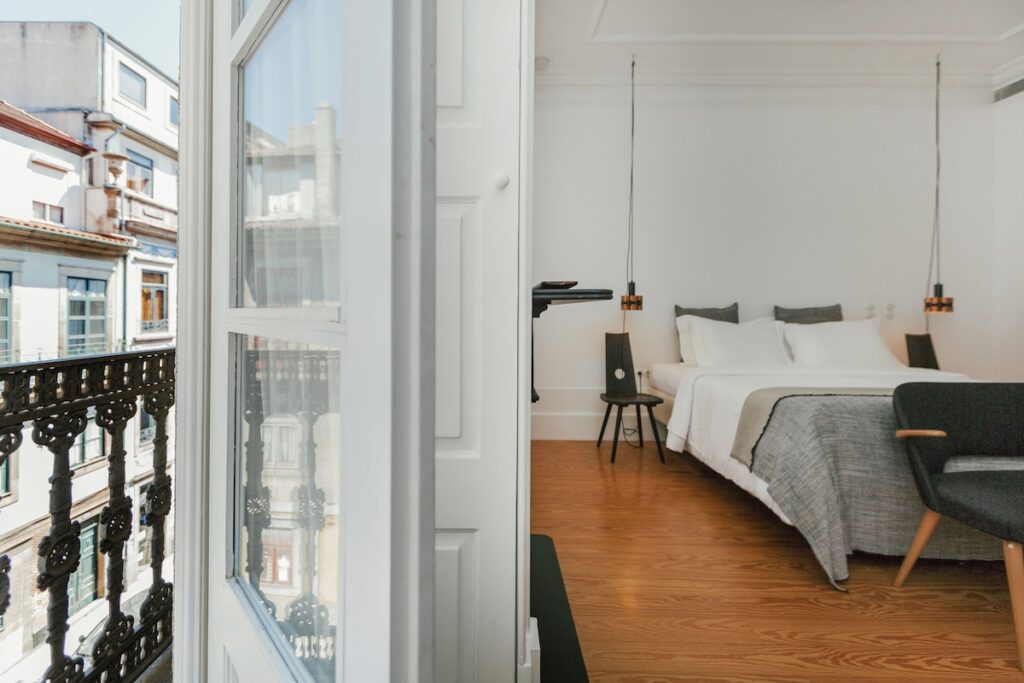 Vista de suíte de hotel de charme no Porto a partir de varanda com guarda-corpo de ferro, exibindo quarto com piso de madeira e cama minimalista.