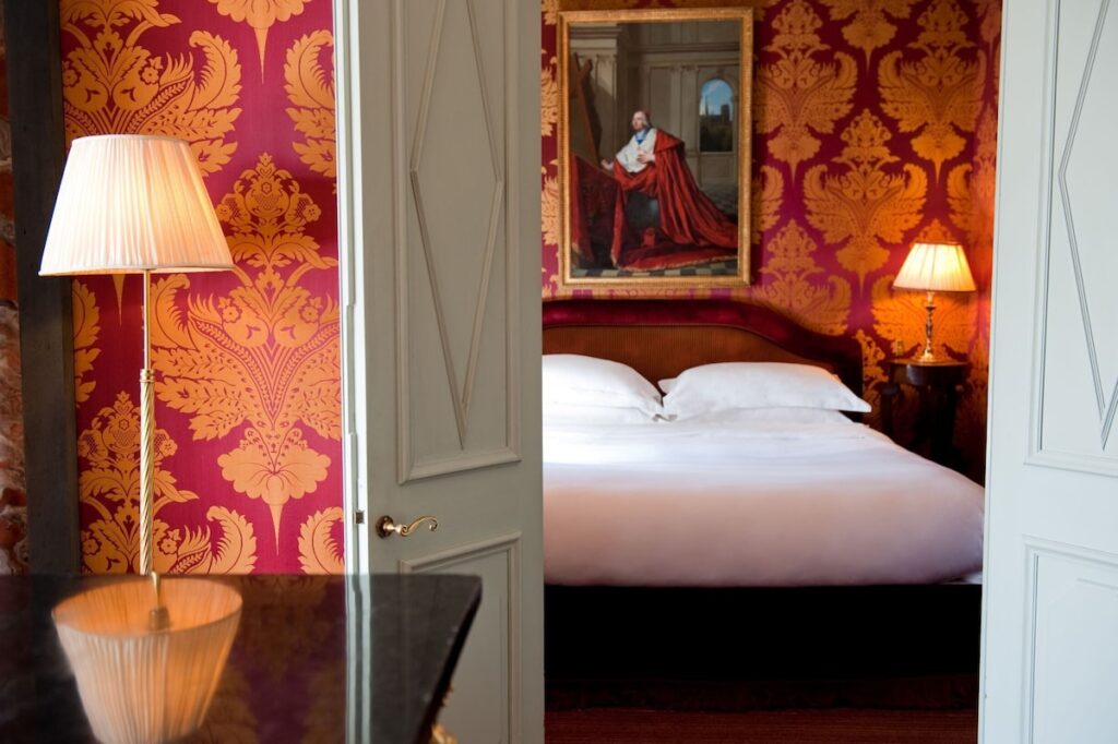 Quarto elegante do L'Hotel, destaque entre hotéis boutique em Paris, visto através de portas semiabertas, com papel de parede adamascado vermelho e dourado.