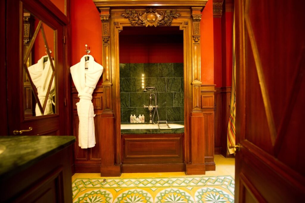 Banheiro de luxo do L'Hotel, ícone entre hotéis boutique em Paris, com portal de madeira entalhada, banheira em mármore verde e piso decorado.
