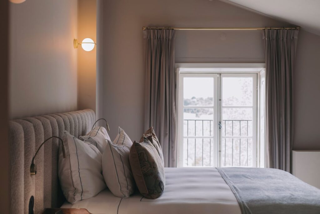 Quarto de hotel com teto inclinado aconchegante, cabeceira de gomos e iluminação quente de um charmoso hotel boutique no Porto.