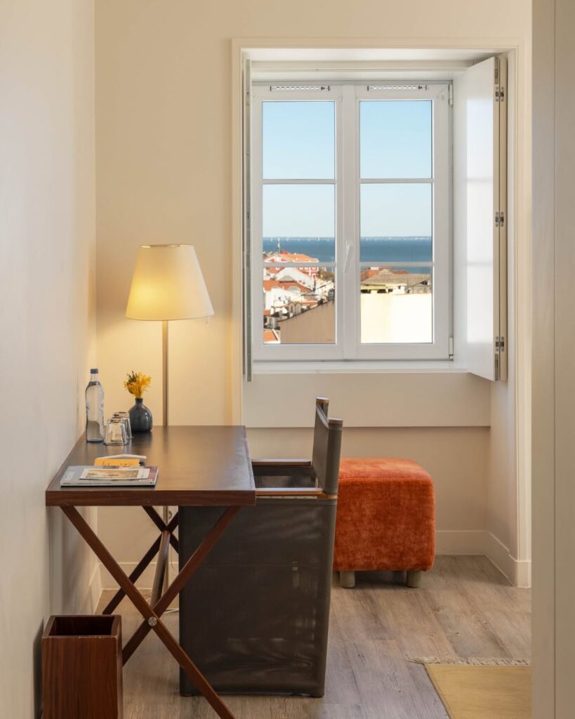 Cantinho de trabalho em suíte do Memmo Alfama, um dos hoteis boutique em lisboa, com mesa de madeira, pufe terracota e janela com vista de rio.