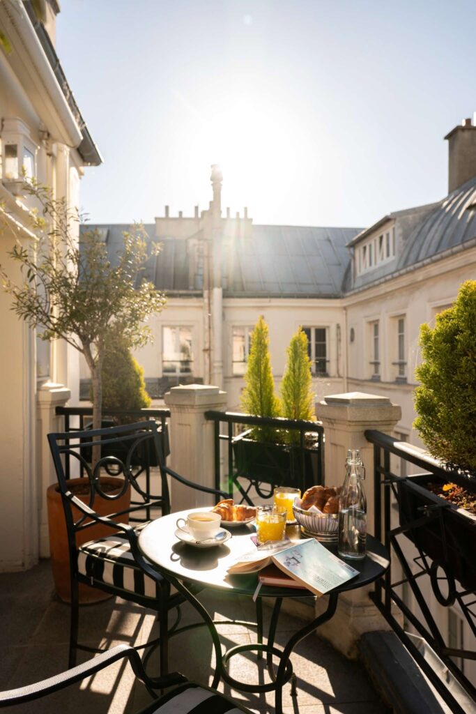 Café da manhã na varanda do L'Hotel, entre charmosos hotéis boutique em Paris, com vista para o pátio interno, mesa de ferro e croissants sob o sol.