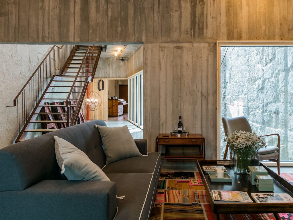 Lounge moderno com sofá cinza, escada de metal e parede de concreto em requintado hotel boutique no Porto. Foto do Armazém Luxury Housing.