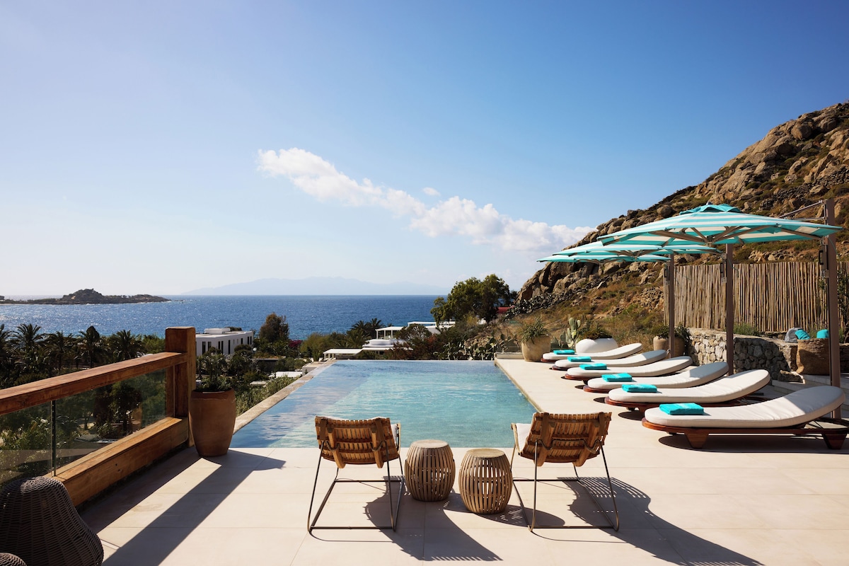 Piscina de borda infinita e espreguiçadeiras com guarda-sóis listrados na Villa do Nammos Hotel, prestigiado resort em Mykonos.