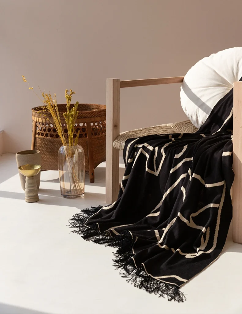 Poltrona de madeira com manta preta de franjas e grafismos, ao lado de cesto de palha, representando decoração aconchegante de inverno.