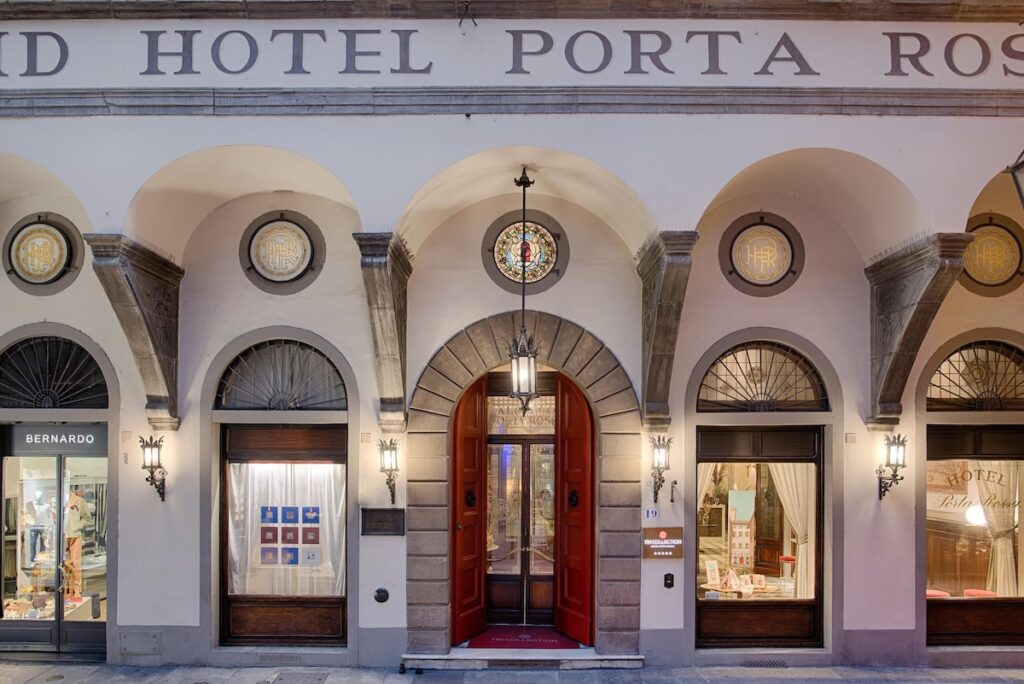 Fachada de hotel histórico em Florença com pórtico em arcos, portas vermelhas e vitrais clássicos do Porta Rossa, combinando herança e design.