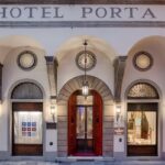 Fachada de hotel hist&oacute;rico em Floren&ccedil;a com p&oacute;rtico em arcos, portas vermelhas e vitrais cl&aacute;ssicos do Porta Rossa, combinando heran&ccedil;a e design.