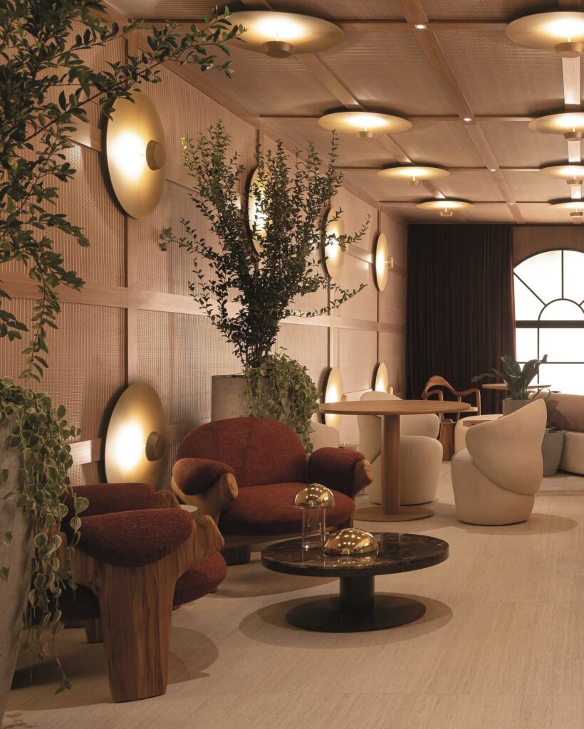 Lounge elegante do Qalb Boutique Café na CasaCor 2026 com poltronas orgânicas vermelhas, painéis de madeira, palha e iluminação cênica dourada.