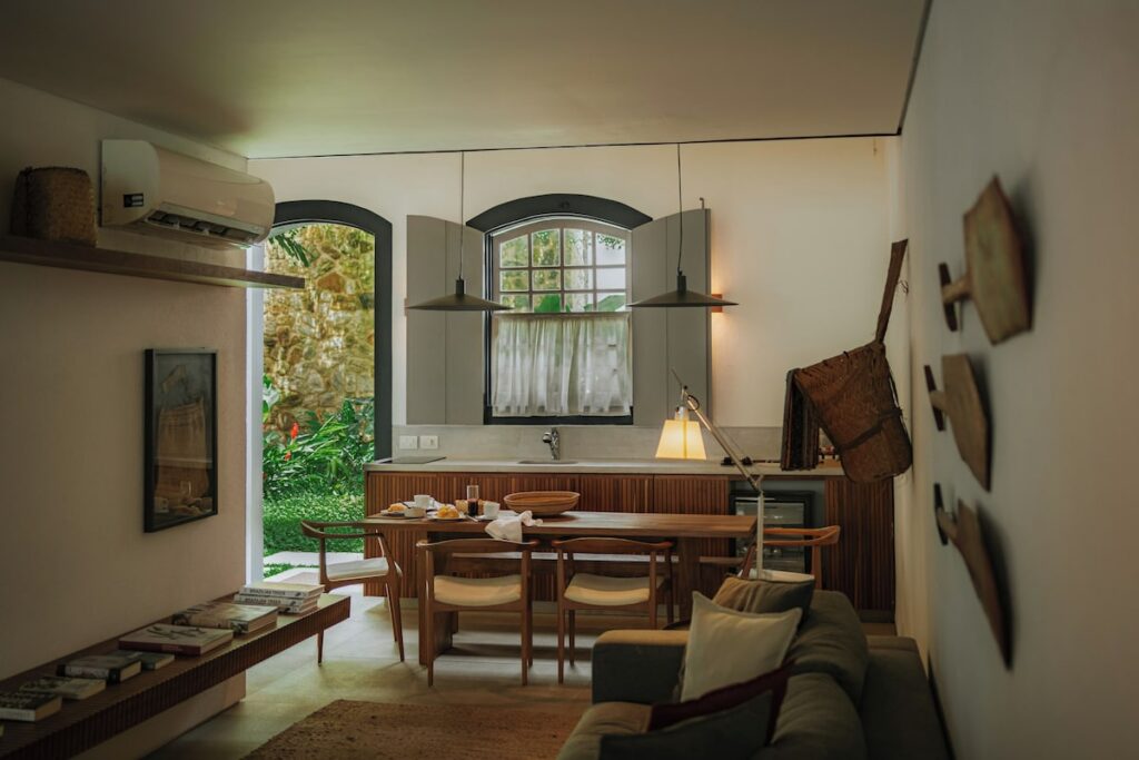 Sala de estar integrada com mesa de jantar de madeira e detalhes coloniais na Pousada Literária de Paraty, com remos decorativos e jardim ao fundo.