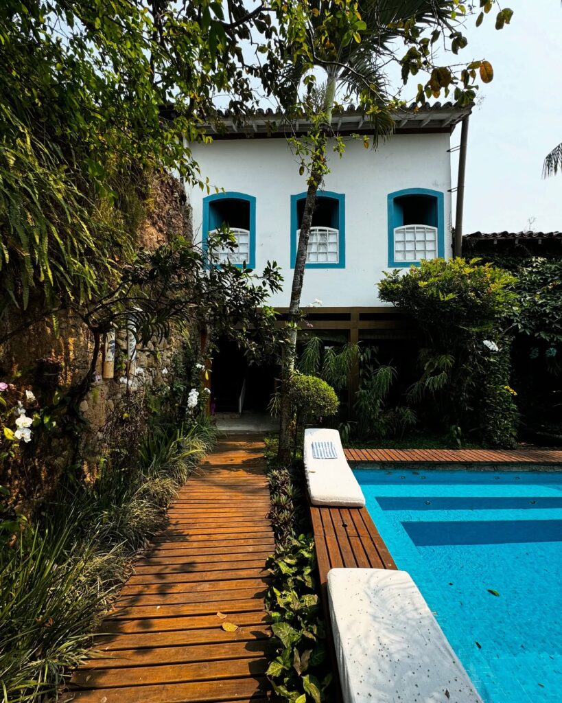 Área externa da Casa Turquesa Maison d'Hôtes com piscina azul, deque de madeira e fachada colonial branca com janelas azul-turquesa e orquídeas.