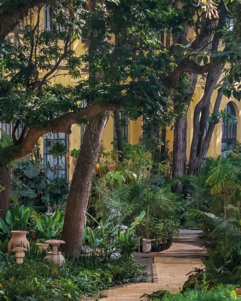 Jardim com caminho em piso cerâmico, vegetação tropical e esculturas de terracota no espaço Da Terra ao Solo Casacor 2026.