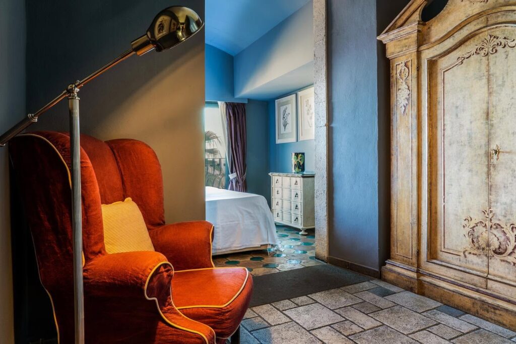 Suíte com poltrona vermelha de veludo, armário clássico entalhado e paredes azul-acinzentadas no Hotel Villa Carlotta Taormina na Sicília.