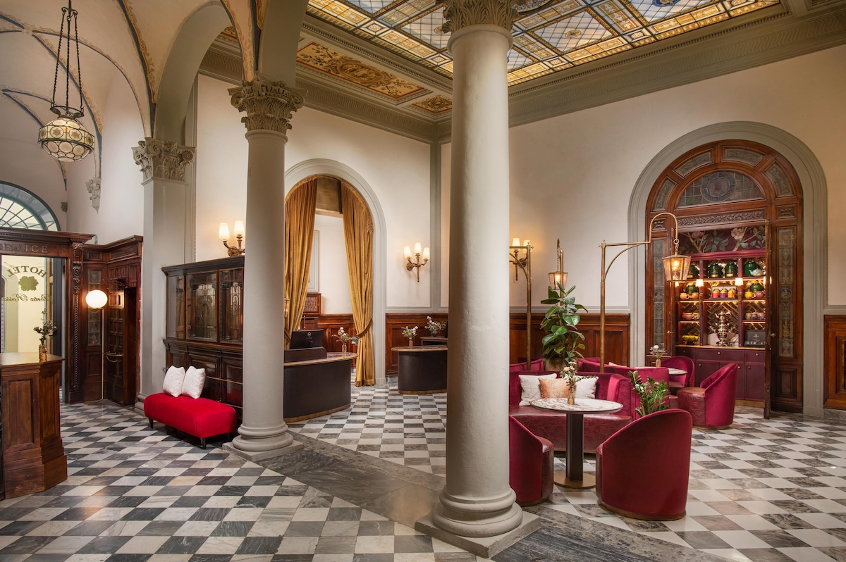 Saguão de hotel de luxo Porta Rossa com piso de mármore xadrez, colunas clássicas, móveis em veludo carmesim e teto ornamentado com afrescos e vitrais.