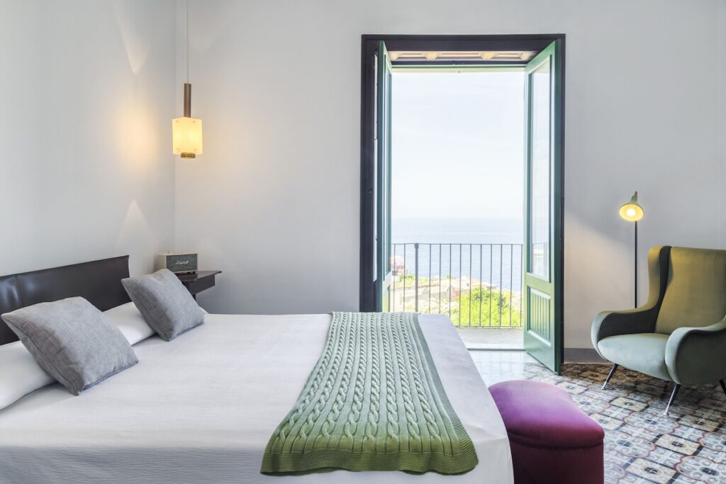 Suíte com piso de ladrilho hidráulico, poltrona oliva e porta aberta para o mar na varanda do Hotel Signum Salina na Sicília.