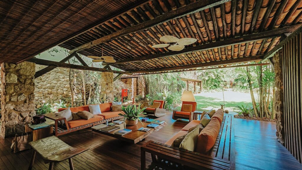 Lounge integrado com sofás terracota, teto de bambu e colunas de pedra na recepção da Pousada Literária de Paraty, com jardim tropical ao fundo.