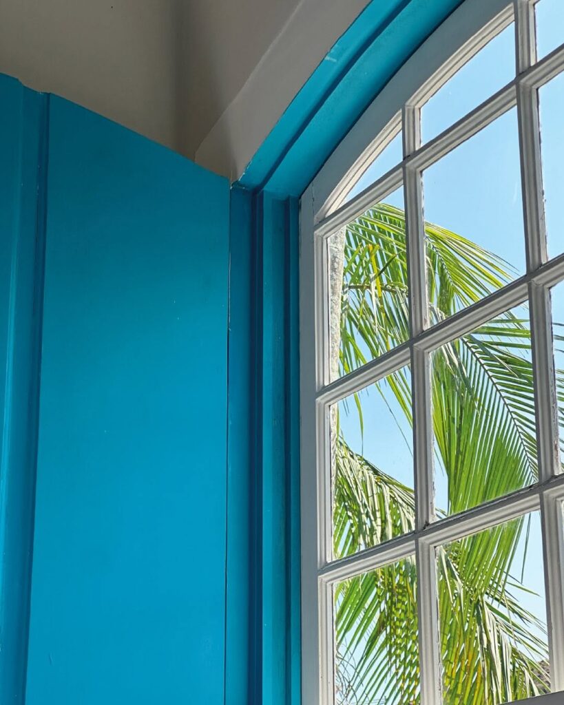 Detalhe de janela colonial de madeira na Casa Turquesa Maison d'Hôtes, com caixilhos brancos e batente azul-turquesa com folha de palmeira ao fundo.