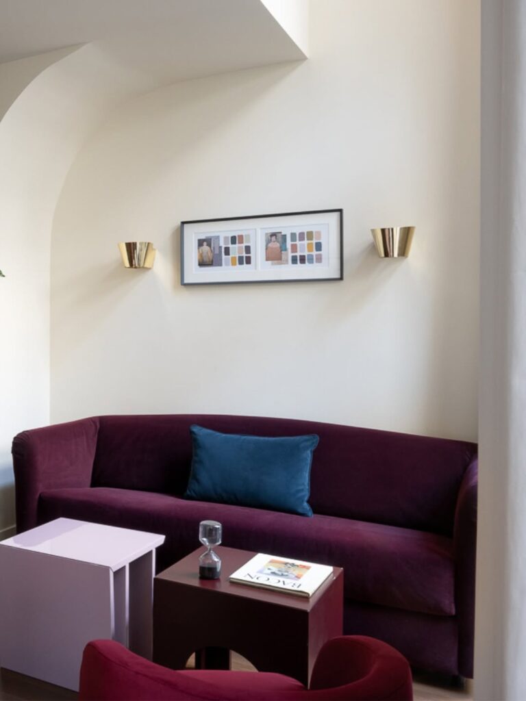 Sofá de veludo roxo com almofada azul e mesas laqueadas coloridas sob arandelas douradas em sala minimalista do Palazzo Talìa.