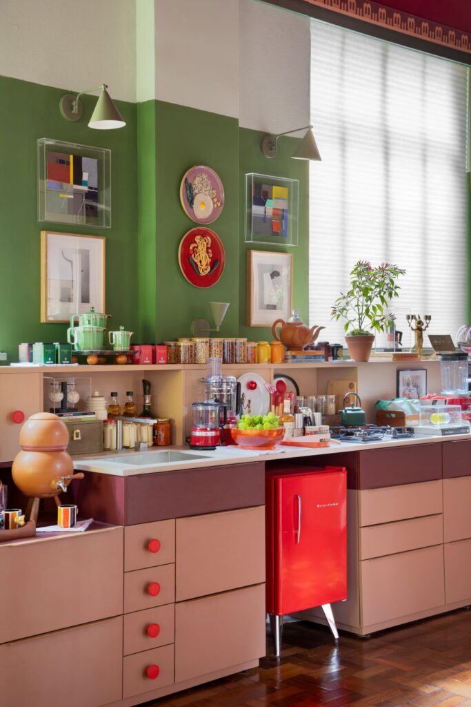 Bancada de cozinha na Casacor São Paulo 2026 com frigobar vermelho retrô Brastemp, marcenaria rosa e parede verde-oliva com pratos artísticos.