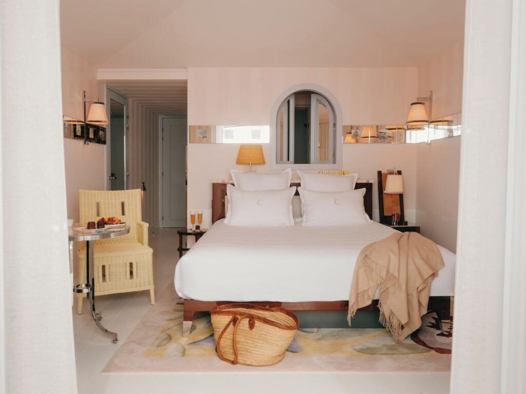 Quarto de casal clássico projetado por Philippe Starck, exibindo tons pastel, cadeira de vime amarela e cabeceira de madeira com espelhos.