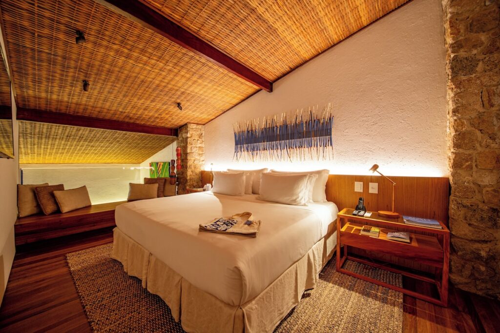 Quarto de casal na Suíte Cajaíba da Pousada Literária de Paraty, com teto inclinado de palha trançada, cabeceira iluminada e tapete de sisal.