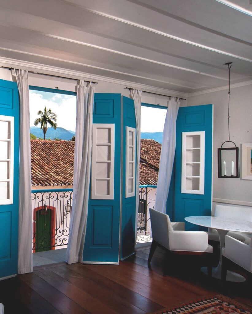 Quarto na Casa Turquesa Maison d'Hôtes com portas coloniais azul-turquesa abertas para sacada, mesa de mármore e cadeiras contemporâneas brancas.