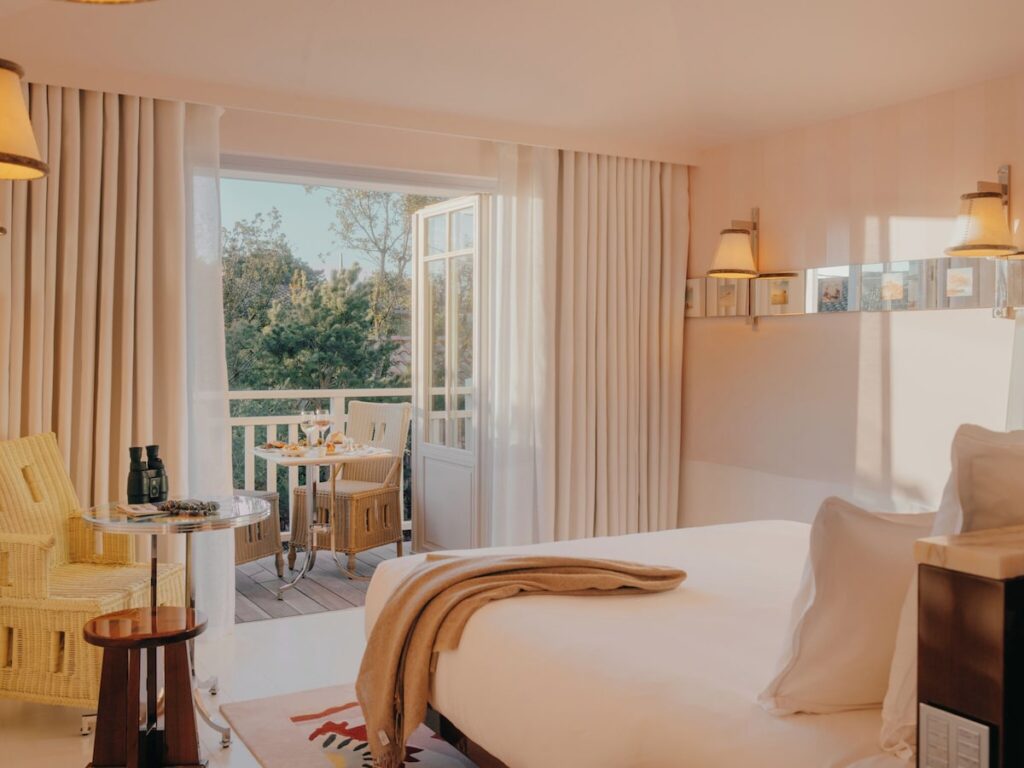 Quarto de hotel luxuoso com sacada aberta para a mata de pinheiros, integrando a natureza local com a narrativa espacial interna de tons claros.