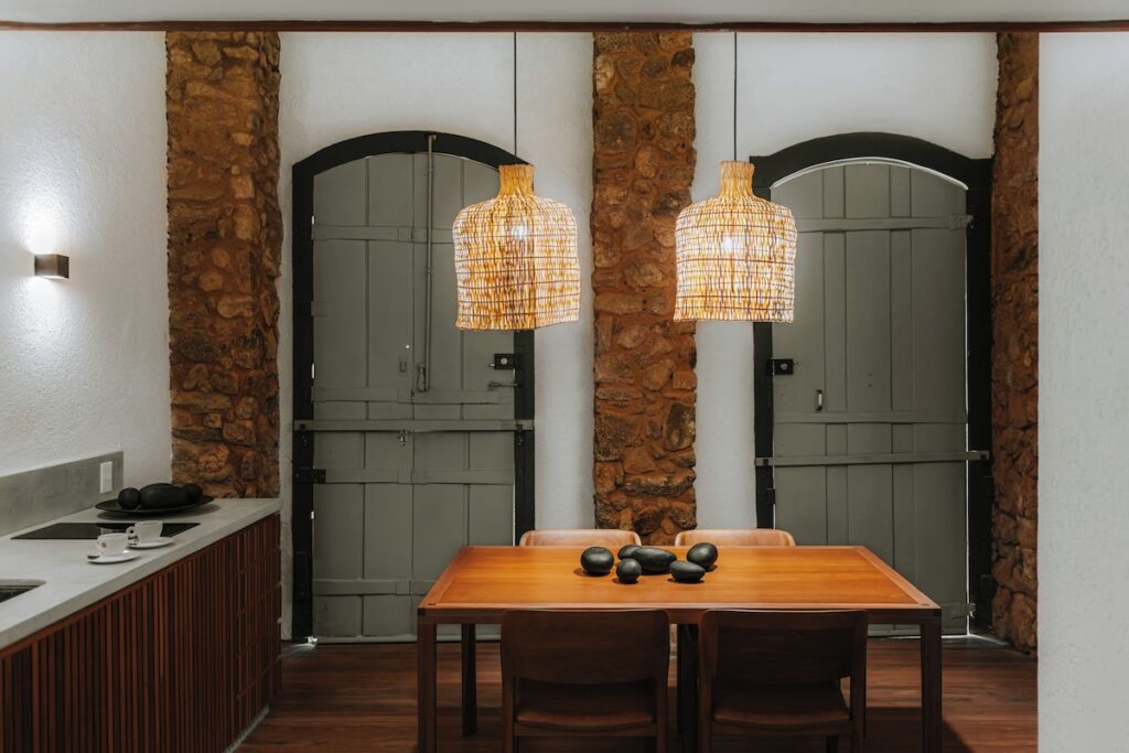 Sala de jantar na Suíte Cajaíba da Pousada Literária de Paraty, com mesa de madeira, luminárias de vime trançado e portas coloniais verde-sálvia.