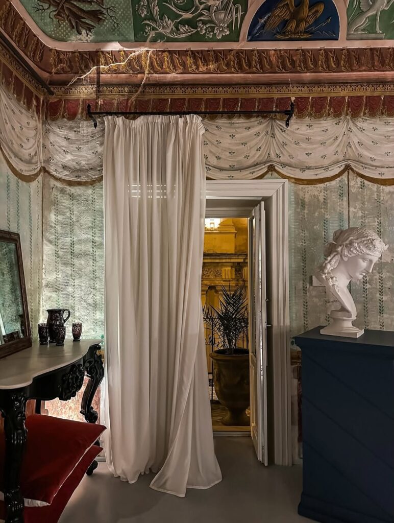 Salão barroco com teto afrescado, busto de gesso clássico, console rococó e cortina branca no Q92 Noto Hotel na Sicília.