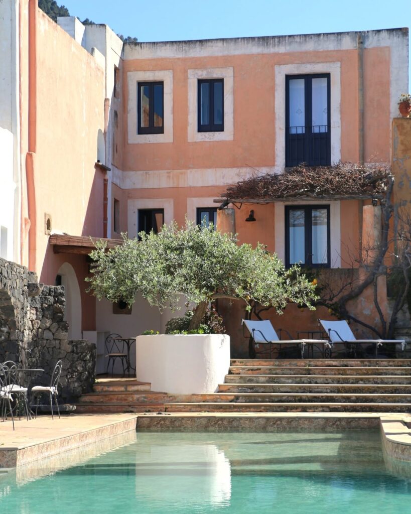 Área externa com piscina turquesa, oliveira central e fachada em tom pêssego no Hotel Signum Salina na Sicília.