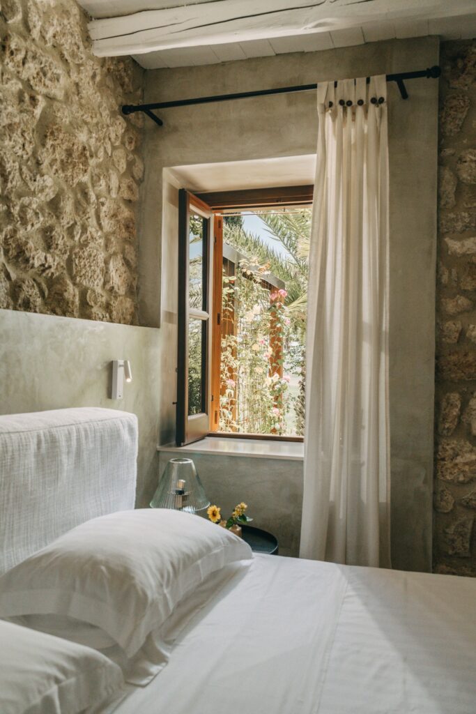 Quarto minimalista com cabeceira branca, parede de pedra rústica e janela aberta para jardim de rosas na Masseria Susafa na Sicília.