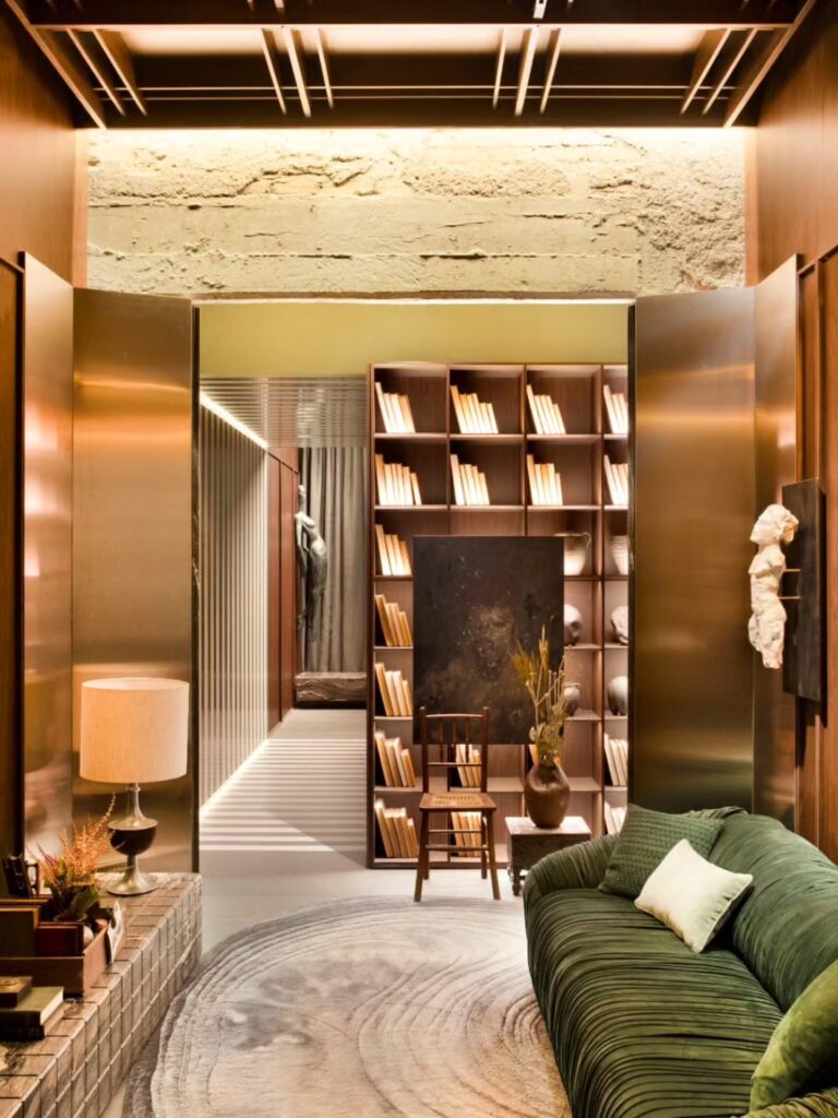 Lounge com sofá de veludo verde, portas de bronze e estante de livros no espaço Banheiro da Saudade Casacor 2026.