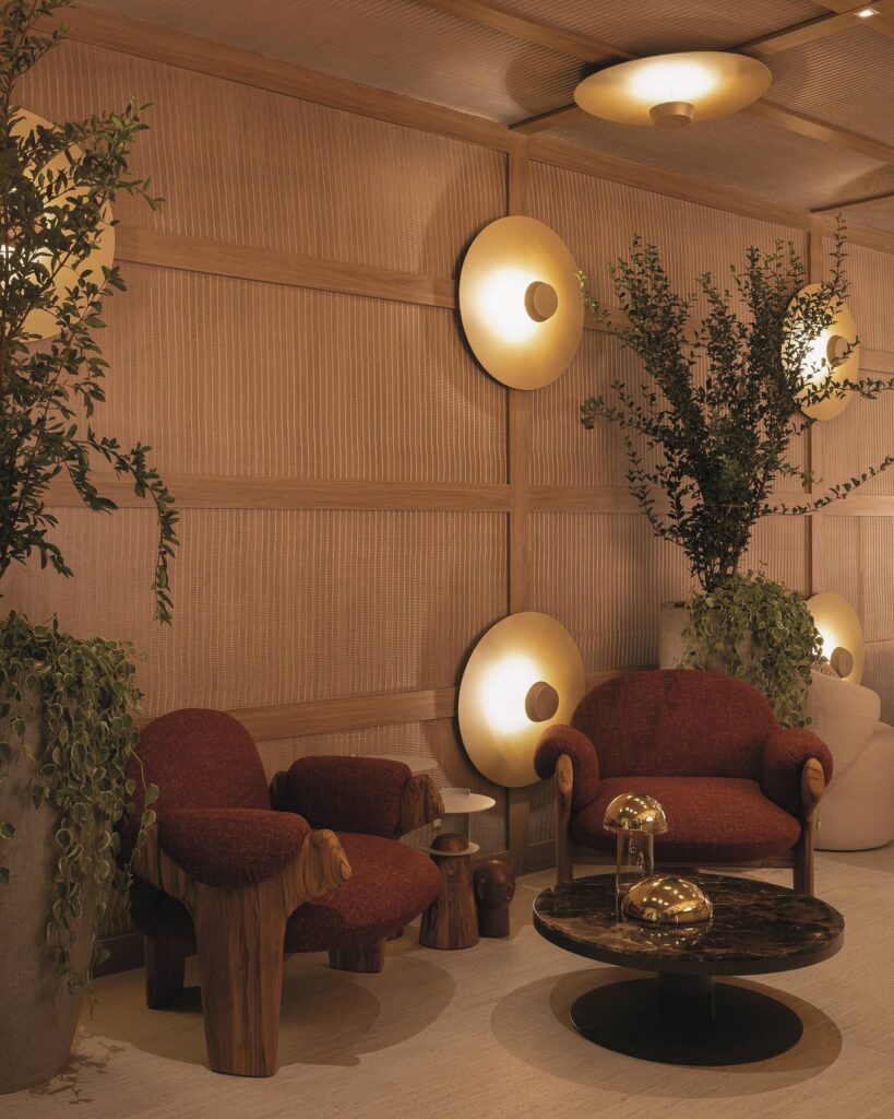Poltronas de madeira e estofado vermelho no lounge do Qalb Boutique Café na CasaCor 2026 com arandelas douradas em painel de palha.