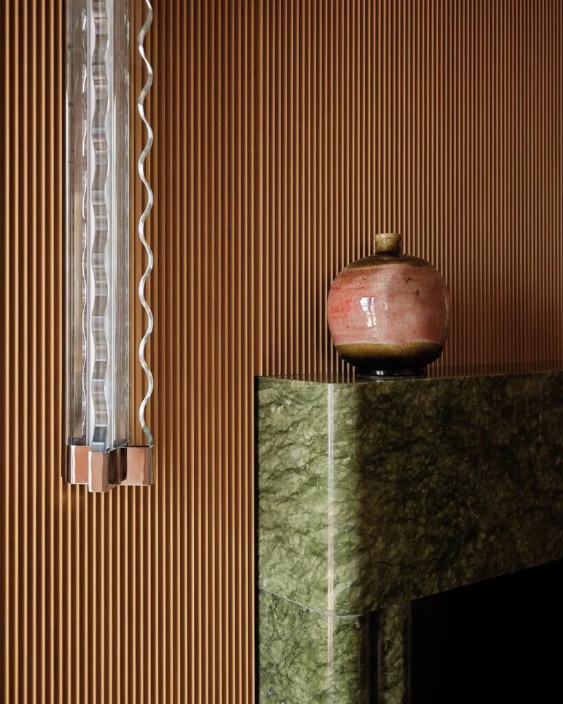 Detalhe de arandela de vidro ondulado sobre parede ripada de madeira e lareira de mármore verde no hotel Palazzo Talìa.
