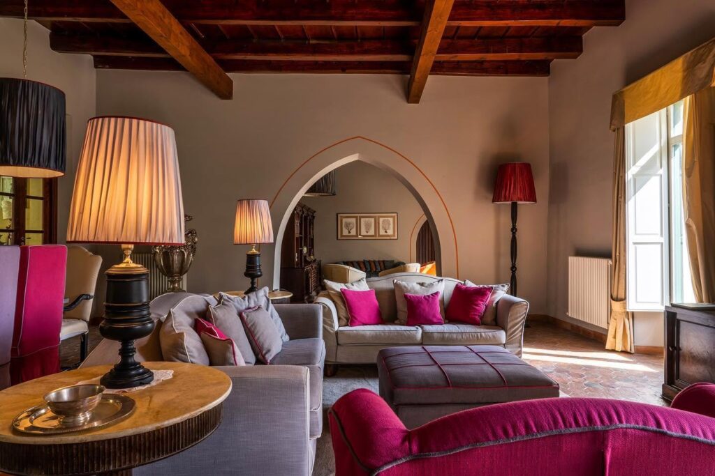 Salão com teto de vigas de madeira, arco ogival gótico, sofás cinza e fúcsia na Villa Teatro Greco Historic House do Hotel Villa Carlotta Taormina.