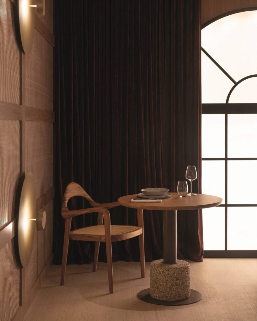 Mesa bistrô e cadeira autoral de madeira no Qalb Boutique Café na CasaCor 2026 com cortina de veludo e janela em arco.