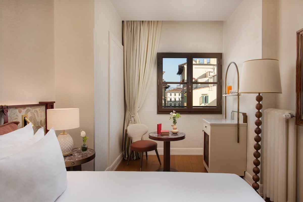 Quarto Solteiro Superior no Porta Rossa com paleta bege clara, luminária escultural de esferas, móvel vintage delicado e janela com vista para Florença.