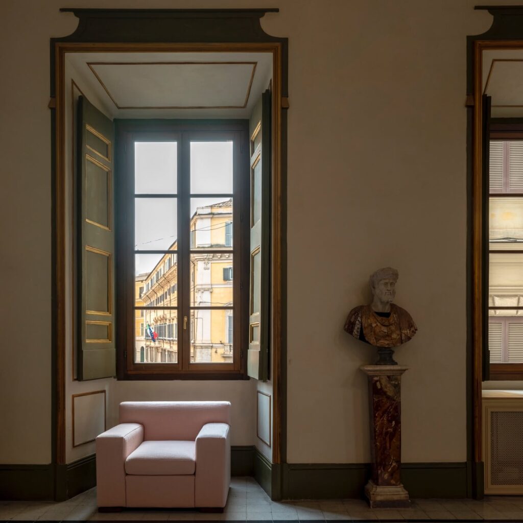 Janela histórica com moldura verde e dourada ao lado de poltrona rosa e busto clássico romano no Palazzo Talìa.