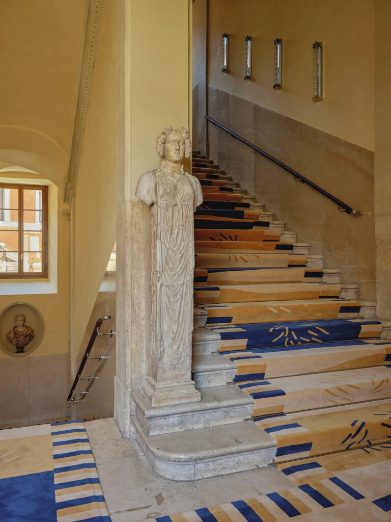 Escadaria histórica com passadeira de carpete geométrico e estátua clássica romana no hotel Palazzo Talìa.