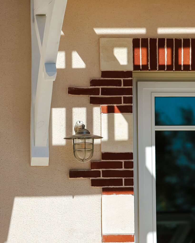 Detalhe de arandela náutica na parede bege com tijolos vermelhos e sombras projetadas do pergolado no hôtel villa colette.