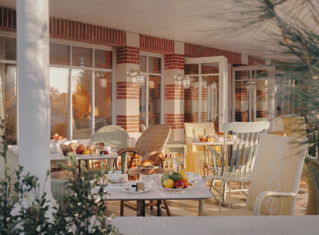 Pátio externo do restaurante do hôtel villa colette com mesas de café da manhã e cadeiras de vime sob a varanda com colunas brancas.