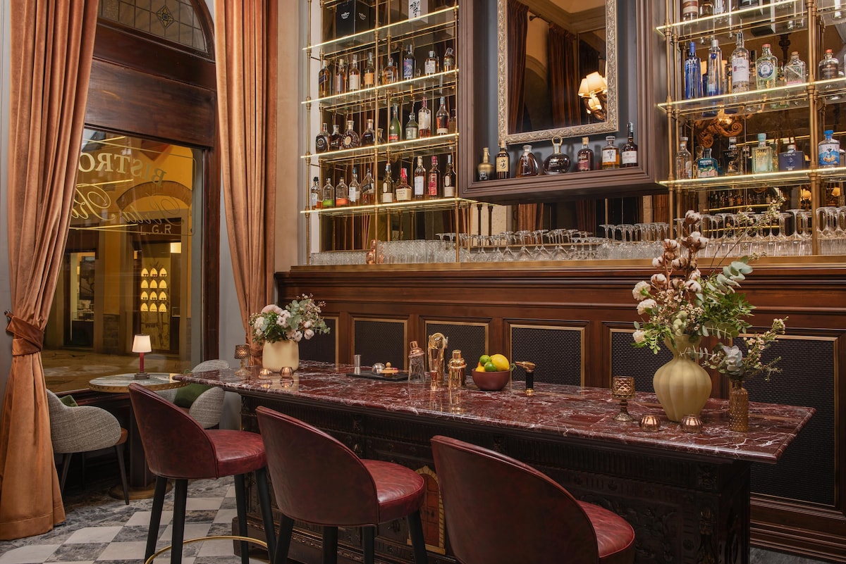 Bar elegante no Porta Rossa com imponente balcão de mármore vermelho, banquetas de couro carmesim, prateleiras de metal finas e cortinas de veludo ocre.