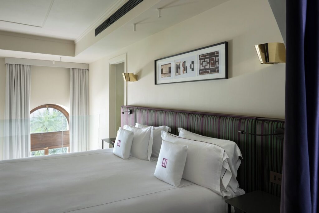Cama de casal com cabeceira listrada na Suíte Premium do Palazzo Talìa, com mezanino e vista para janela arqueada.