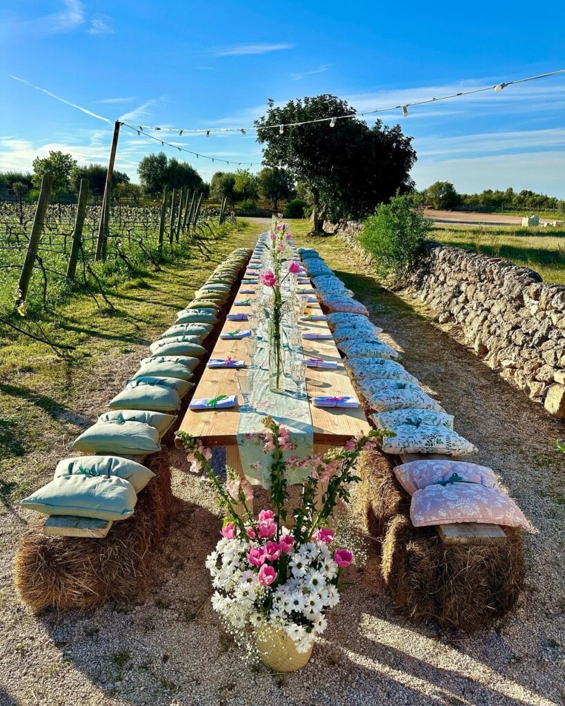 Mesa comunitária em vinhedo com fardos de palha e arranjos florais na Masseria Montenapoleone.